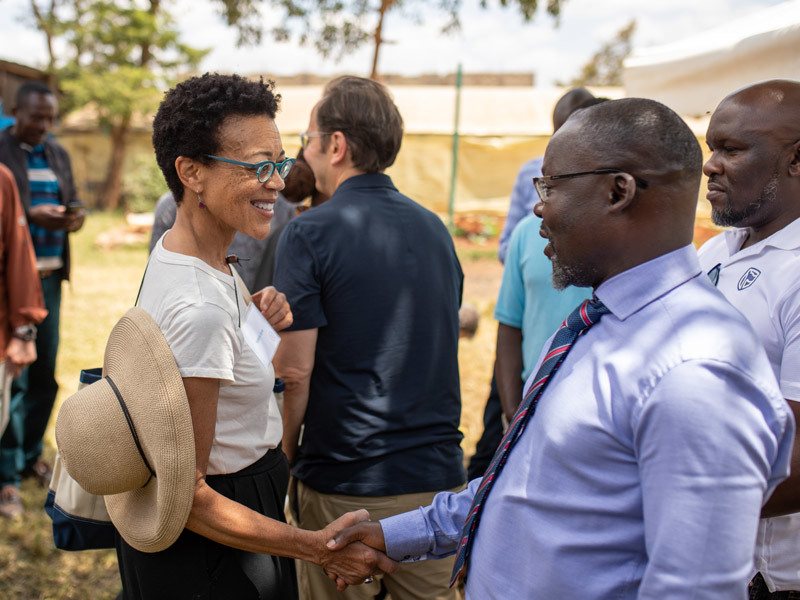 two people shaking hands outside and smiling. the woman is wearing a white tshirt and a straw hat hanging around her neck and blue glasses. the man is wearing a light blue collared shirt and a tie and glasses.